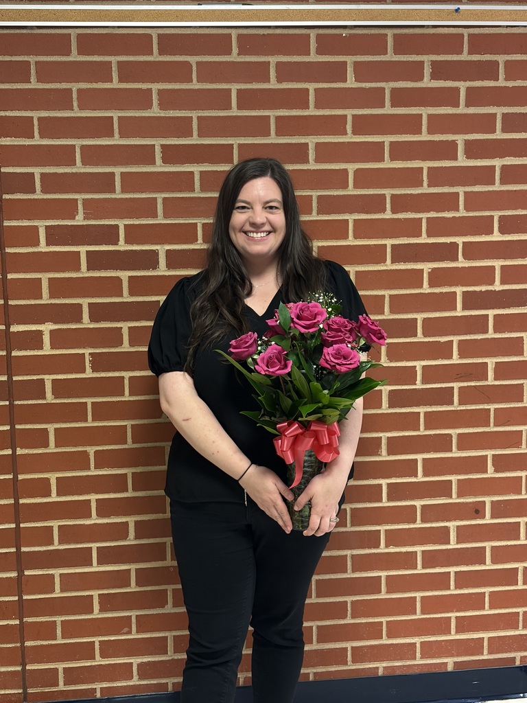 A smiling woman with long dark hair stands in front of a red brick wall, holding a bouquet of bright pink roses tied with a red ribbon. She is wearing a black outfit and faces the camera, appearing proud and happy.