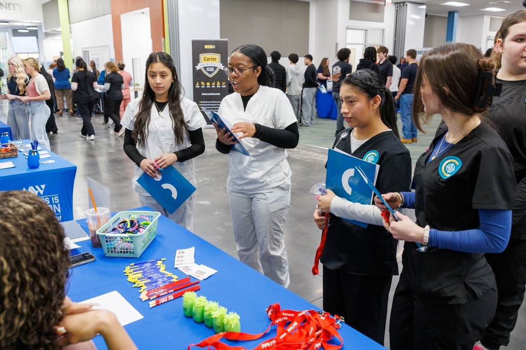 Students in healthcare uniforms speak with a recruiter at a table during a senior job fair.