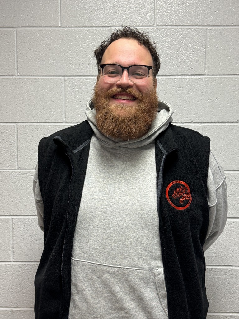 A smiling man with glasses and a full beard stands in front of a light-colored brick wall. He is wearing a gray hoodie under a black vest with a red logo. He appears friendly and approachable, with his hands behind his back.
