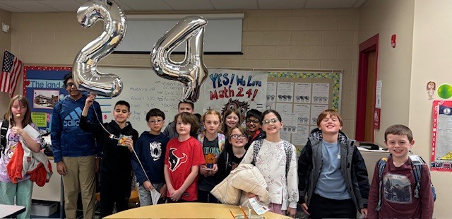 A group of students poses in a classroom with large silver balloons shaped like the numbers “2” and “4.” Some students wear backpacks and jackets, and a “YES! We Love Math 24!” sign is visible behind them on the wall.
