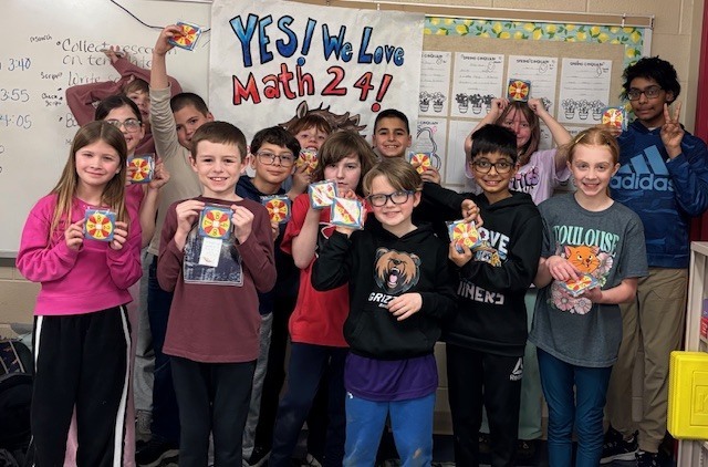 A group of elementary students stands in a classroom in front of a whiteboard and a colorful sign that reads “YES! We Love Math 24!” The students smile and hold up small square cards with colorful designs, appearing proud of their work.