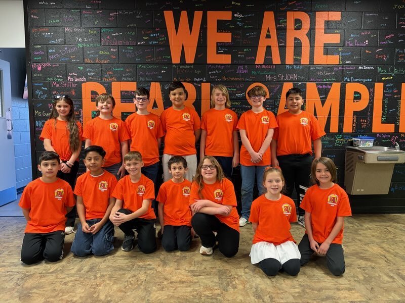 A group of elementary students in matching orange shirts stand and kneel in rows in front of a decorated wall that says “WE ARE PEARL SAMPLE,” smiling and ready for their Math 24 competition.