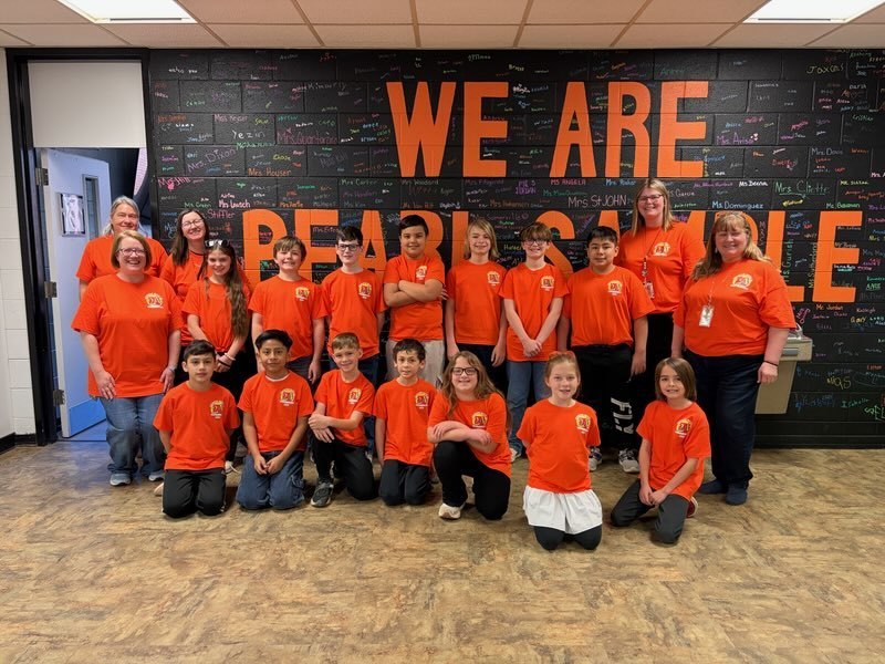 A group of elementary students and several staff members wearing bright orange shirts pose together in front of a black wall covered with colorful writing and large orange letters that read “WE ARE PEARL SAMPLE.”