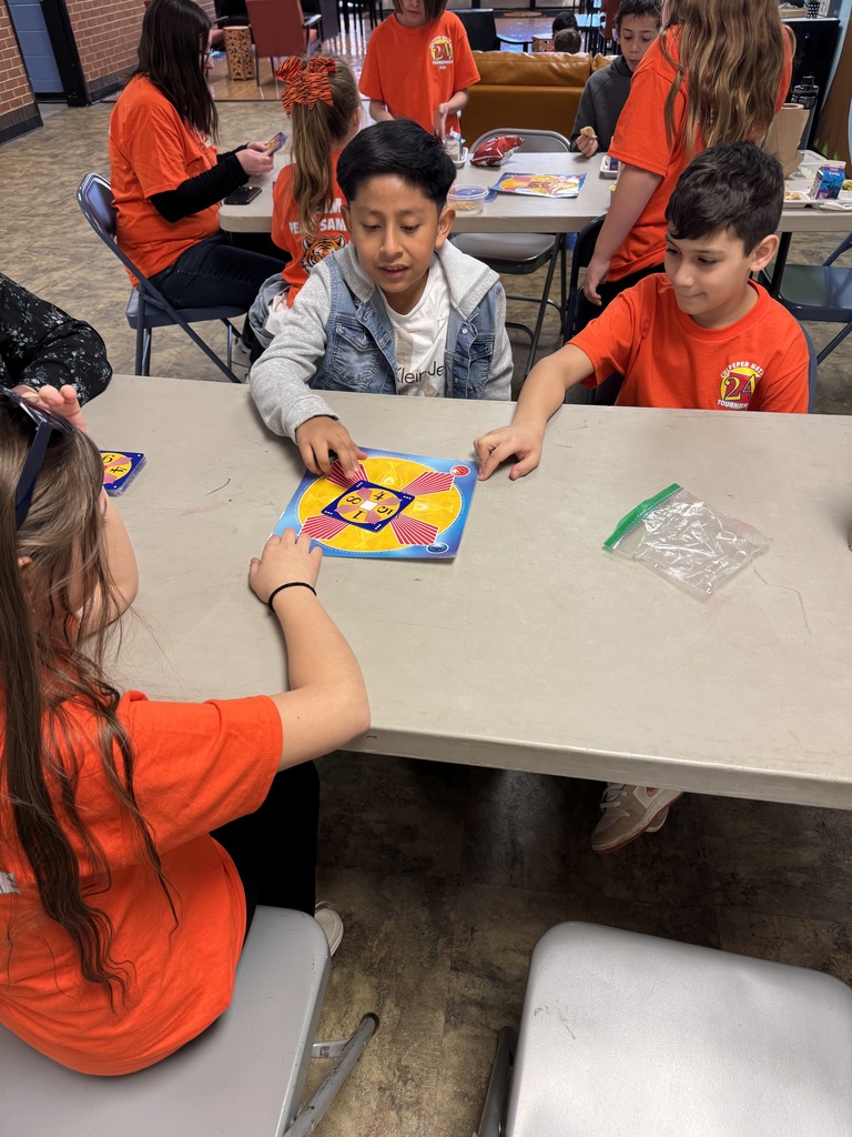 Several students in orange shirts sit around a table playing a Math 24 card game, focusing on solving problems together as they prepare for their upcoming competition.