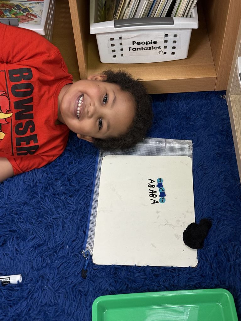 A student smiles while lying on a rug next to a whiteboard showing a simple repeating pattern labeled with letters.