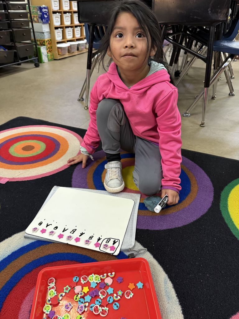 A student in a pink hoodie sits on a colorful rug, using small shaped manipulatives to create a pattern and label it on a whiteboard.