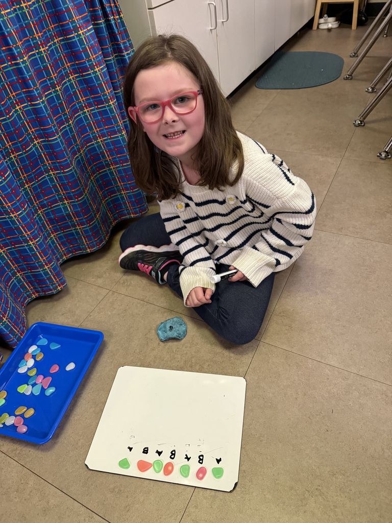 A student with glasses sits on the floor using colorful stones to build a repeating pattern and identify the pattern core on a whiteboard.