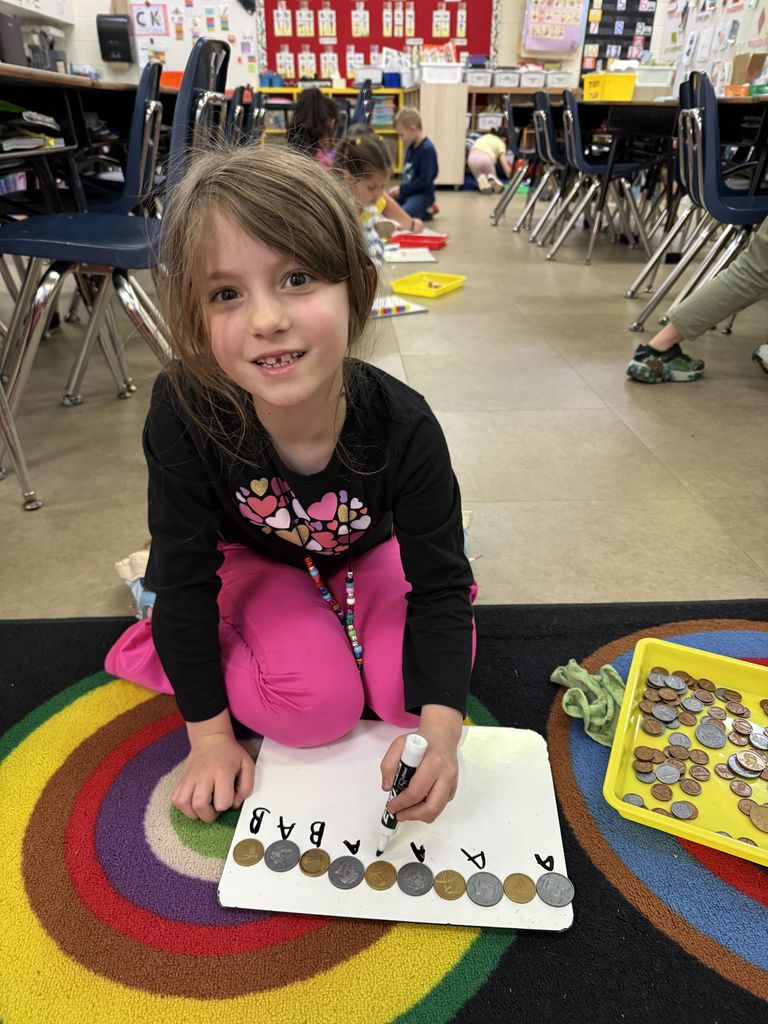 A student sits on a rug with coins, arranging them into a pattern and labeling the sequence with letters on a whiteboard.