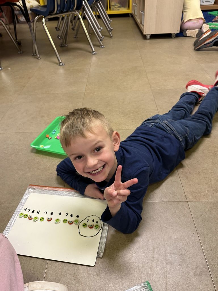 A student is on the floor smiling and holding up a peace sign while showing a completed repeating pattern made with small objects and labeled on a whiteboard.