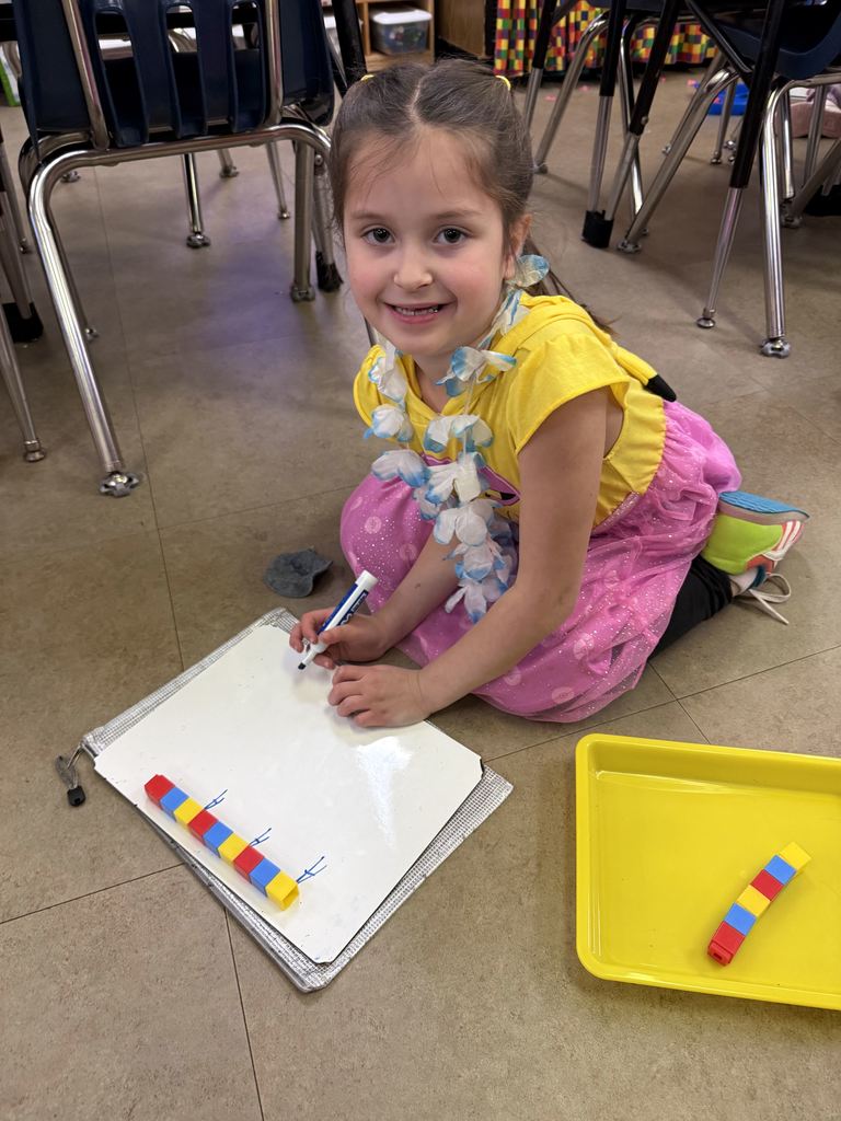 A smiling first-grade student kneels on the floor, building a colorful cube pattern and drawing matching marks on a whiteboard.