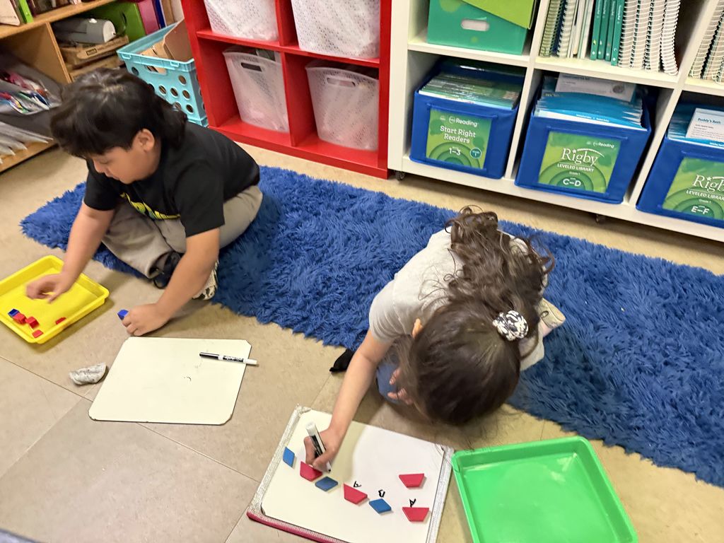 Two students work independently on a rug, arranging pattern blocks and recording their patterns with letters on whiteboards.