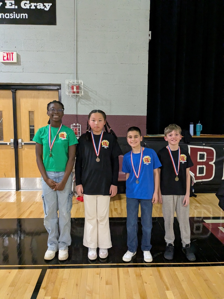 Four elementary students stand side by side in a gym wearing medals around their necks, smiling after being recognized as winners in a school competition.