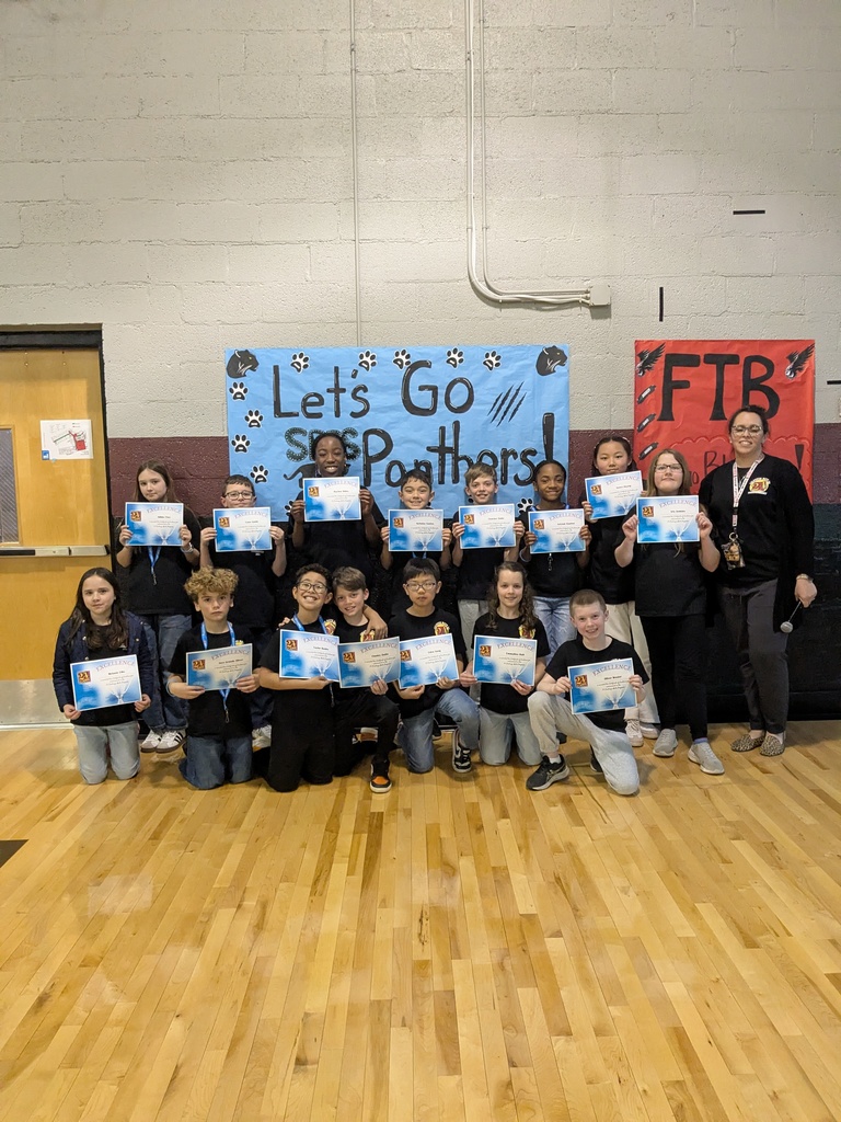 A large group of elementary students and a teacher pose in a gym holding certificates of excellence, standing in front of a “Let’s Go Panthers” sign.