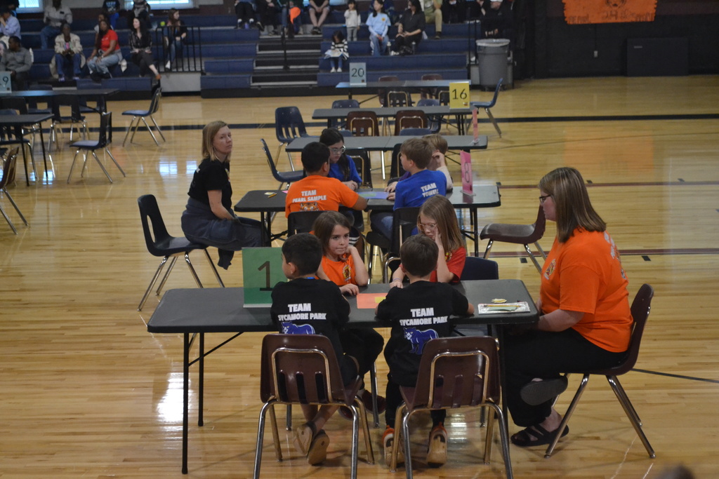 Several teams of elementary students sit at numbered tables across a school gym floor, working together on a competition while teachers supervise and spectators sit in the bleachers.