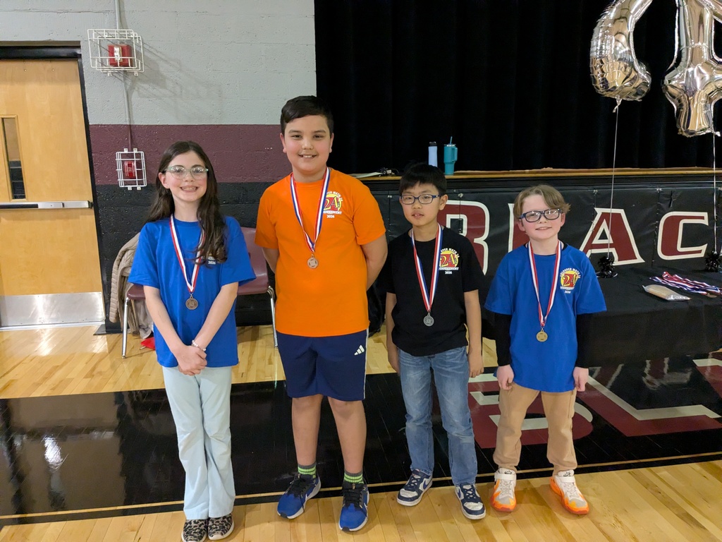 Four elementary students wearing medals stand in a row in a gym, smiling proudly after receiving awards for a school competition.