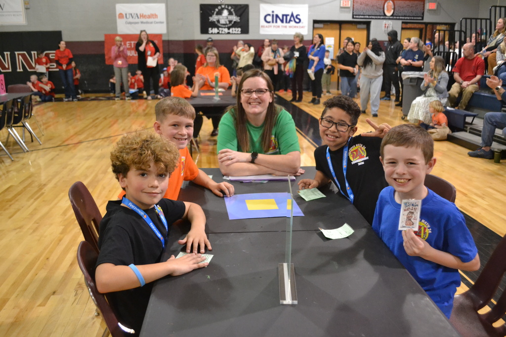 A smiling teacher sits with four elementary students at a table during a school competition in a gymnasium, while a crowd of students and adults watch and clap in the background.