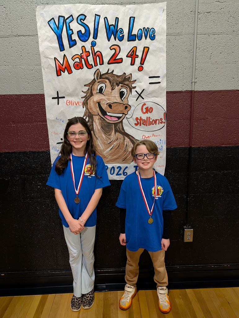 Two elementary students wearing blue “Math 24 Tournament” shirts and medals stand smiling in front of a large hand-drawn poster that reads “YES! We Love Math 24!” with a cartoon horse mascot and “Go Stallions!” written beside it.