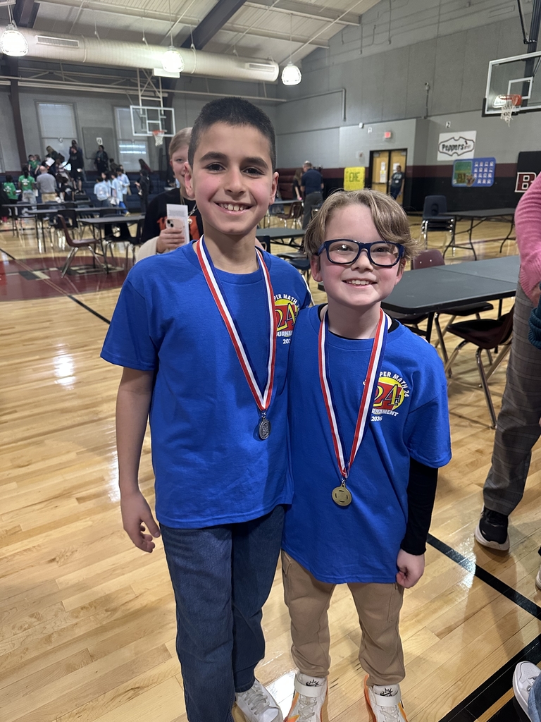 Two elementary students wearing blue Math 24 Tournament shirts and medals stand side by side in a school gym, smiling at the camera with tables and other participants visible in the background.