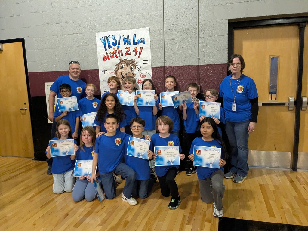 A group of elementary students in matching blue Math 24 Tournament shirts pose together in a gym holding certificates, with two adults standing on either side and a “YES! We Love Math 24!” poster behind them.