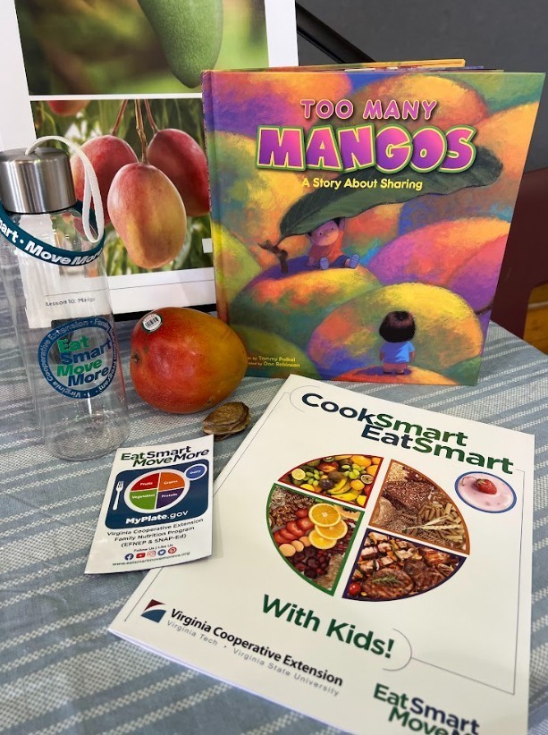 Colorful display of nutrition education materials on a table. A children’s book titled “Too Many Mangos: A Story About Sharing” stands upright next to a real mango and a reusable water bottle with an “Eat Smart, Move More” logo. In front, a brochure titled “Cook Smart, Eat Smart With Kids!” from Virginia Cooperative Extension shows a plate divided into food groups. A small MyPlate-style card and a poster with images of mangoes are visible in the background.