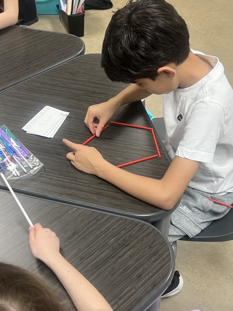 A student leans over a desk, carefully positioning red sticks to form a polygon. A small stack of index cards and a bag of colorful sticks sit nearby.