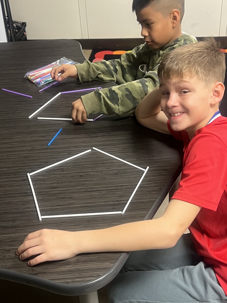 Two students build a polygon using white sticks on a desk. One student smiles at the camera while the other focuses on placing the sticks into shape.