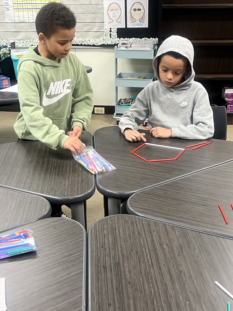 Two elementary students sit at desks working together. One student takes colorful sticks from a bag while the other arranges red and white sticks into a polygon shape on the table.