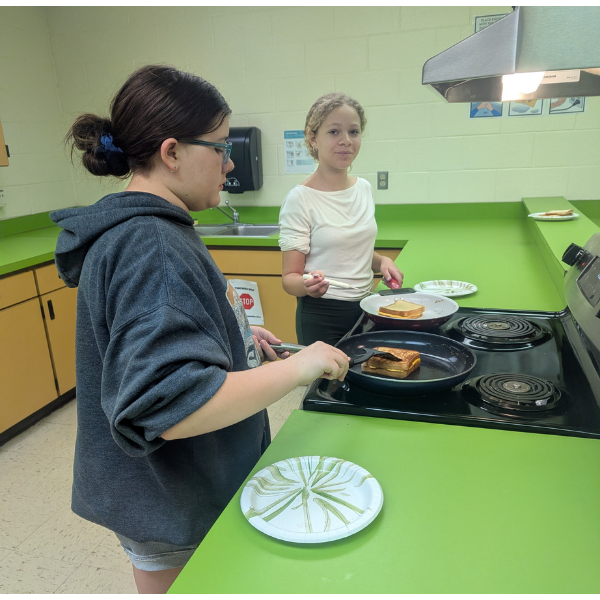 Two students are cooking grilled cheese sandwiches on a stovetop in a classroom kitchen. One student uses a spatula to flip a sandwich in a frying pan while another holds a plate with a finished sandwich. Paper plates and cooking supplies are visible on the bright green countertops, and a sink and cabinets are in the background.