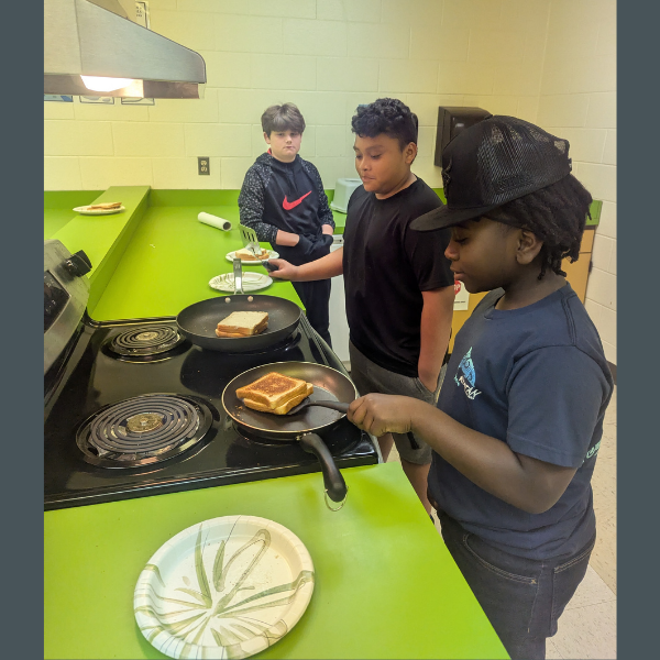 Three students are preparing grilled cheese sandwiches at a stovetop in a classroom kitchen. One student flips a sandwich in a frying pan while another holds a plate and a third watches nearby. The space features bright green countertops, paper plates, and kitchen equipment, creating a hands-on learning environment.