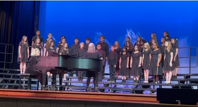 A school choir of about two dozen middle or high school students stands on tiered risers on a stage, wearing coordinated black concert attire. A pianist sits at a grand piano to the left, accompanying the group. The stage is lit with a bright blue backdrop, and the students appear to be singing during a performance.