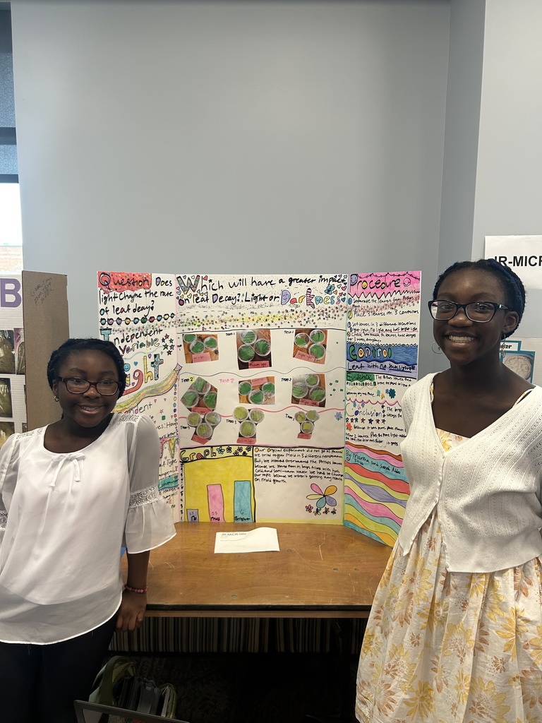 Two students stand smiling beside a colorful tri-fold science fair display about leaf decay. The project explores whether light or darkness has a greater impact on how leaves decay, and includes sections such as question, variables, procedure, observations, graphs, and conclusion, along with photos of the experiment. The display is set on a table in a classroom or event space.