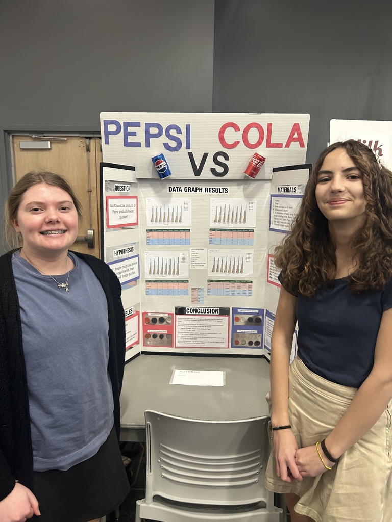 Two students stand smiling beside a tri-fold science fair display titled “Pepsi vs Cola.” The project board includes sections such as question, hypothesis, materials, data graph results, and conclusion, with charts and images displayed. The students are in a classroom or event space, standing in front of a table with a chair and their project presentation.