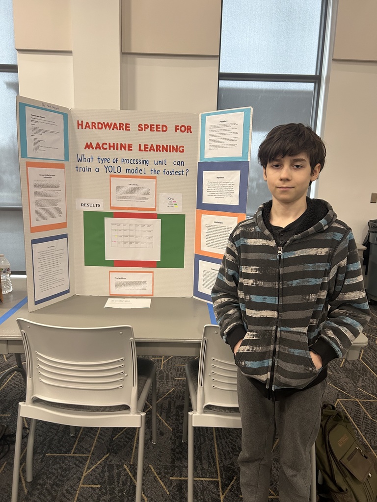 A student stands beside a tri-fold science fair display board titled “Hardware Speed for Machine Learning.” The project asks which type of processing unit can train a YOLO model the fastest. The board includes sections such as background information, procedure, hypothesis, results, limitations, and conclusion, with a chart displayed in the center. The student is wearing a striped hoodie and standing in a classroom or event space with chairs and large windows in the background.