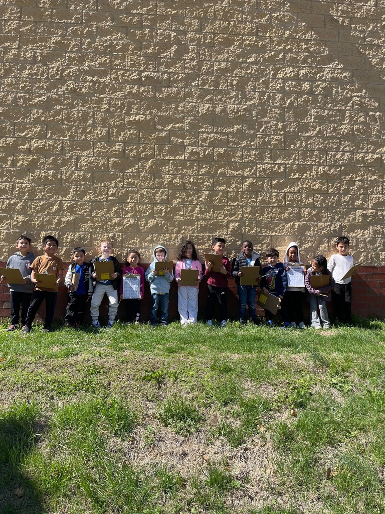 A group of young students stands in a line outside against a tan brick wall, holding clipboards. They are on a grassy area in bright sunlight, smiling and posing together.