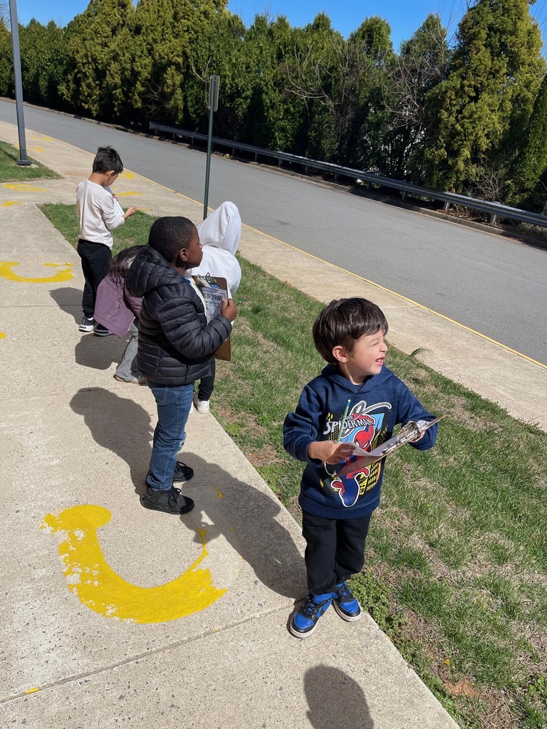 Students stand along a sidewalk near a road, holding clipboards and looking around as they search for signs of spring. Yellow painted markings are visible on the pavement, and trees line the background.
