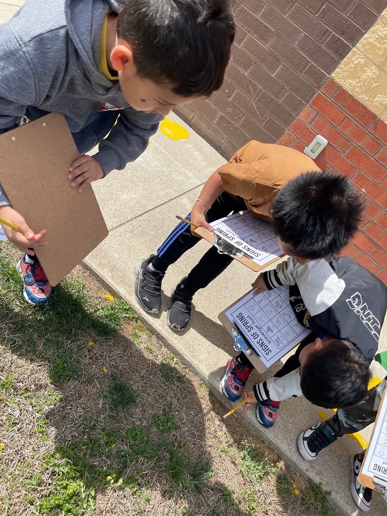 A small group of students gathers around, looking down at a clipboard labeled “Signs of Spring” while writing and observing plants along the sidewalk.
