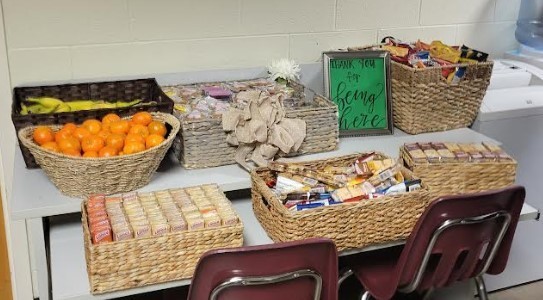A table set up with neatly arranged snack baskets, including oranges, granola bars, crackers, and packaged treats. A small decorative sign reading “Happy you are being here” sits among the baskets, with two chairs placed in front of the table against a light-colored wall.