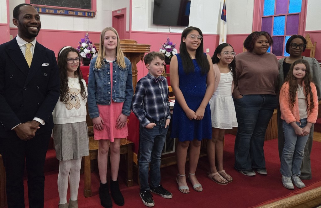 A group of eight elementary and middle school students stand in a row at the front of a church alongside an adult male. The students are dressed in semi-formal attire, including dresses, sweaters, and button-down shirts. Two floral arrangements and a wooden podium are visible behind them, along with pink walls, stained glass windows, and a mounted screen. The group is smiling and facing forward, posing for a recognition photo.