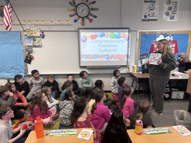 A classroom full of third grade students sit on a colorful carpet facing their teacher, who stands at the front holding up a book. A screen behind her reads “Congratulations Published Authors!!” with balloons, as students look on attentively.