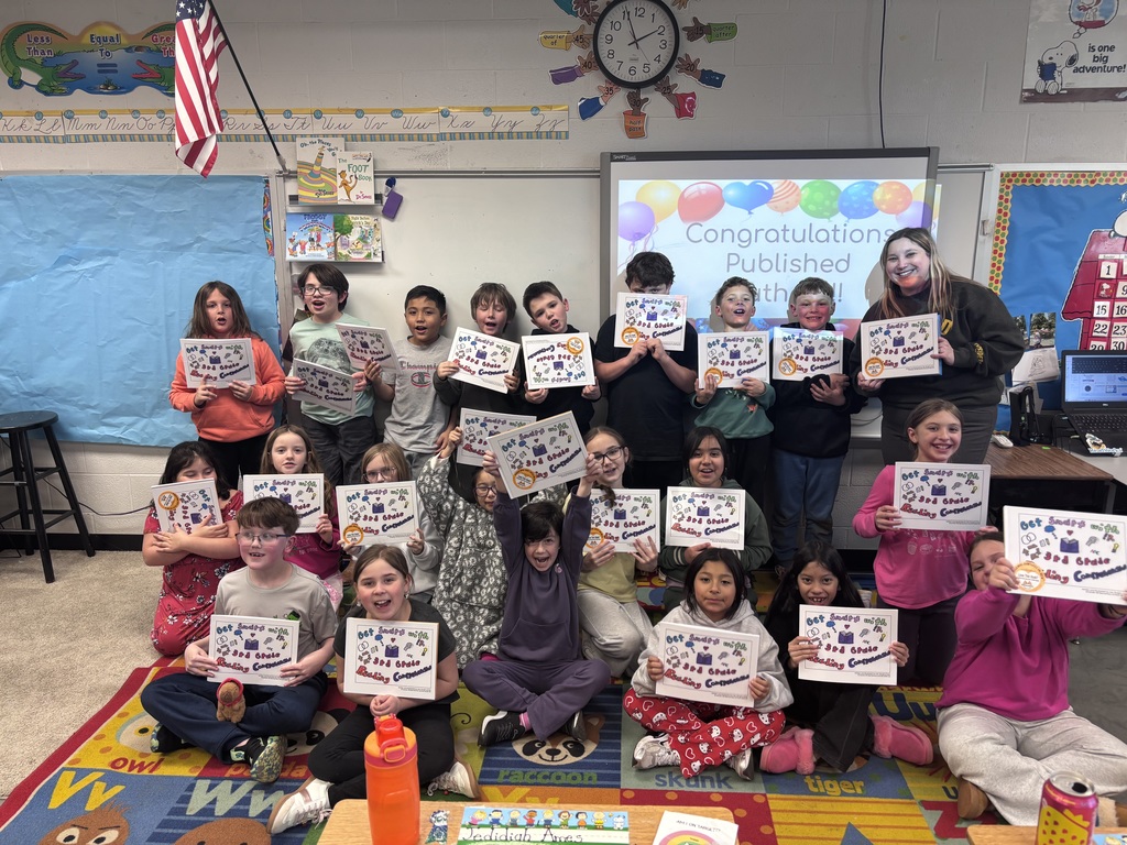 A large group of third grade students and their teacher pose in a classroom holding up copies of their published book. Many students are smiling and displaying their books, celebrating their achievement as published authors.