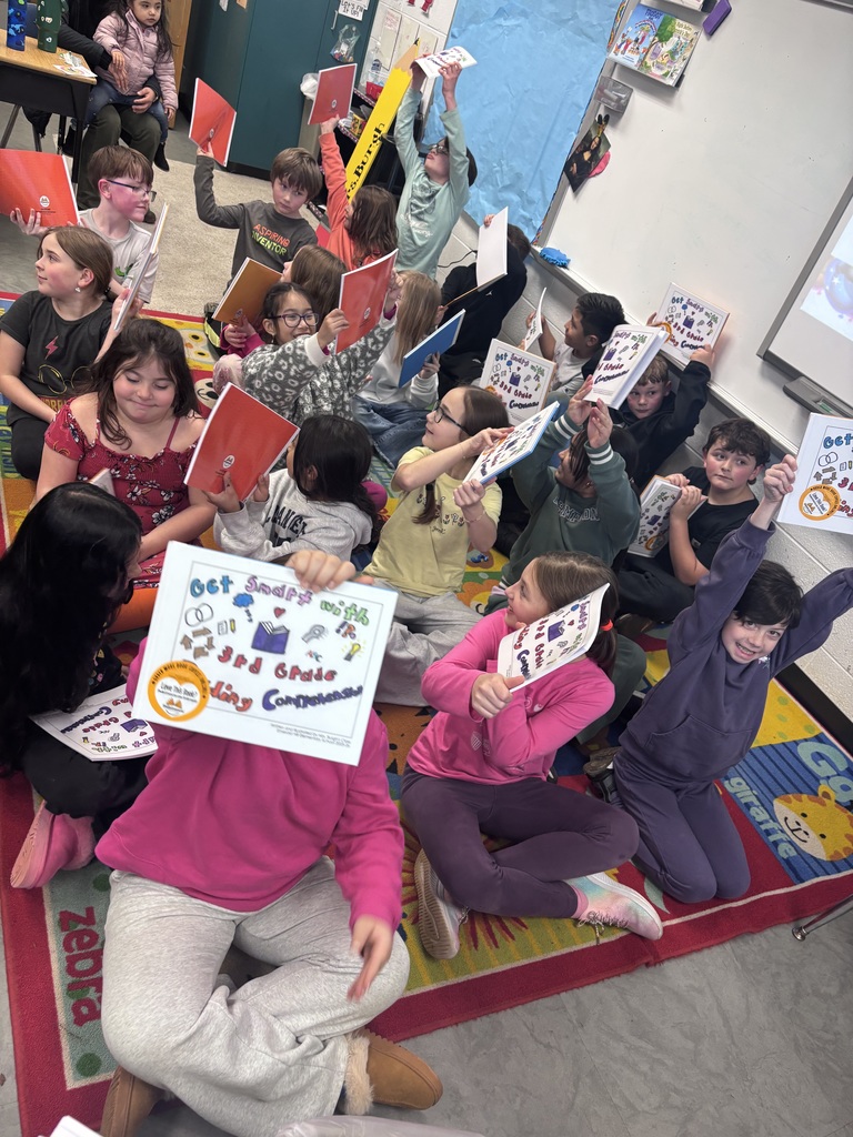 A group of third grade students sit closely together on a classroom carpet, smiling and raising their published books in the air. The students appear excited and proud as they show their work.