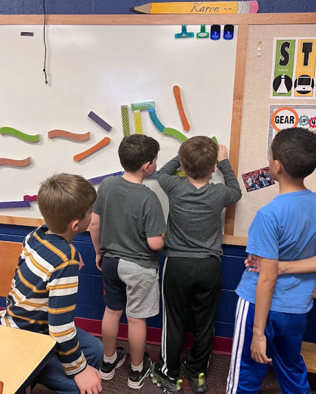 A group of students stands at a whiteboard using colorful flexible strips to create shapes and designs, collaborating side by side.