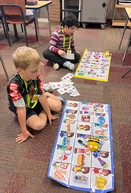 Two students sit on the classroom floor using coding mats and small robotic devices, guiding the robots through a sequence of picture-based commands.