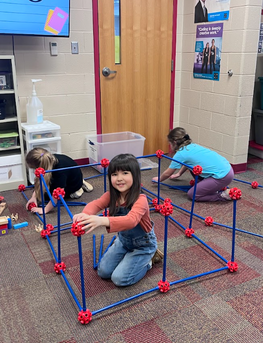 Students sit on the floor assembling multiple cube-shaped structures using blue rods and red connectors, focused on building and connecting pieces.