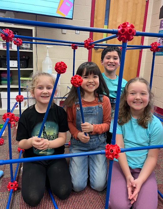 Four smiling students sit and kneel inside a large geometric structure made of blue rods and red connectors, showing off their completed build.