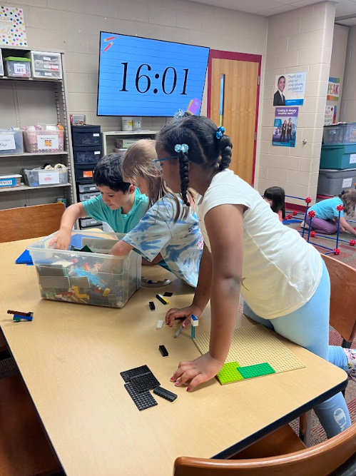 Students gathered around a table in a classroom STEM space, reaching into a bin of building materials and working together to construct with small pieces.
