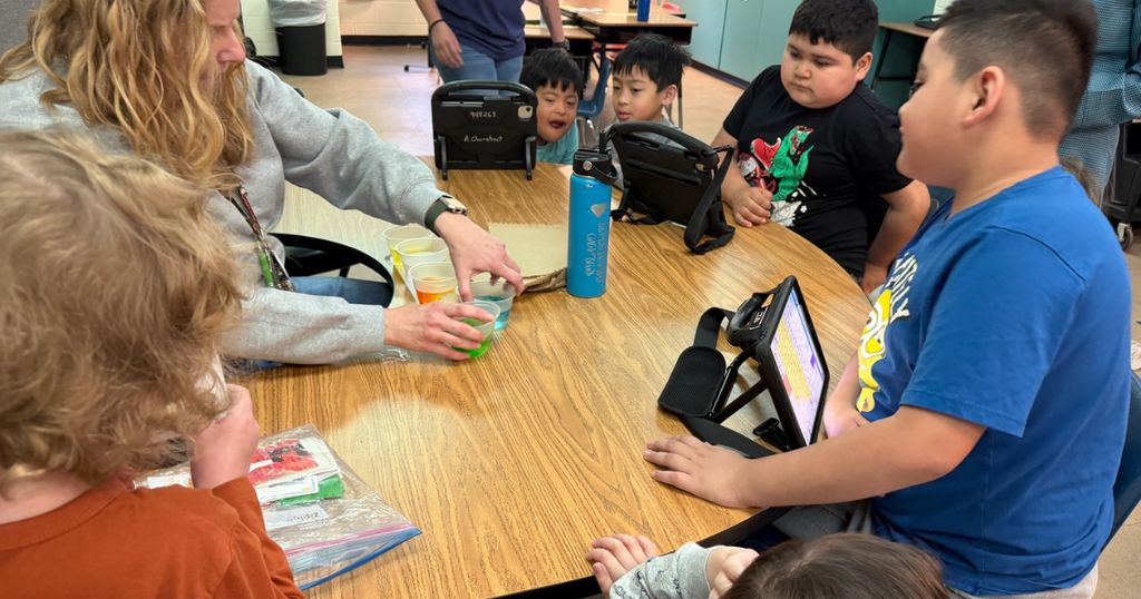 A teacher sits at a classroom table with several students, helping them dip eggs into cups of brightly colored dye while tablets sit nearby.