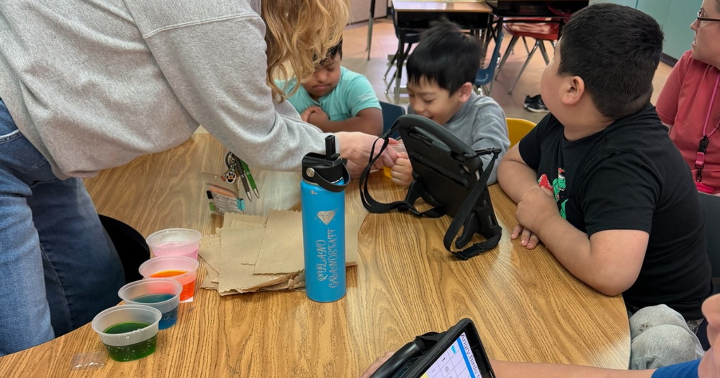 A teacher assists students gathered around a table as they carefully dye eggs in cups of pink, blue, green, and orange liquid.