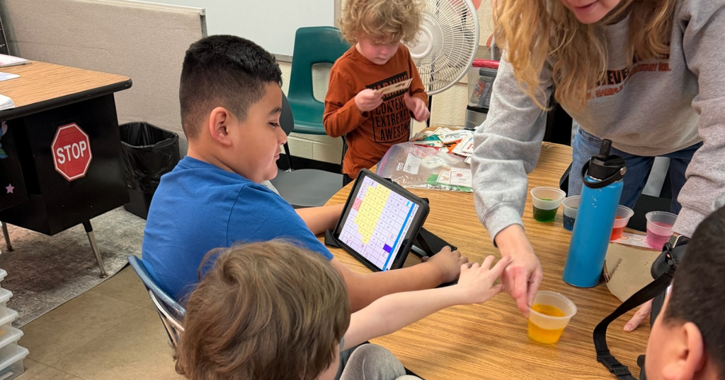 A student watches closely as a teacher guides his hand to dip an egg into yellow dye; other students and colorful cups are visible on the table.