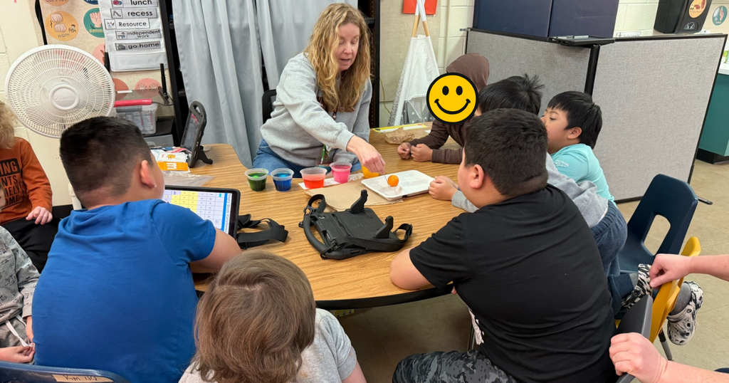 A group of students sits around a table while a teacher demonstrates egg dyeing; several dyed eggs and cups of color are arranged in front of them.